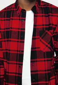 Camicia di flanella a quadri rossi e neri con maniche lunghe, colletto con bottoni e una singola tasca sul lato sinistro del petto; indossata sopra una t-shirt bianca.