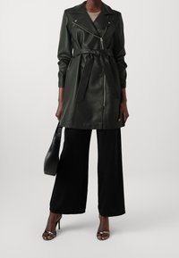 Un abrigo trench de piel sintética de color verde oscuro con cuello con muesca, cierre con cremallera en el frente y cintura ceñida con cinturón. Combinado con pantalones amplios negros y tacones con tiras.