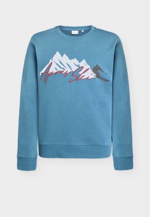 Sweatshirt azul claro com gráfico de montanha e esquiador, "Après Ski" em cursivo vermelho. Decote redondo e punhos e bainha canelados. Material em mistura de algodão.