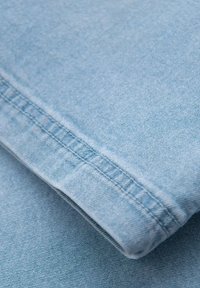 Tissu en denim bleu clair avec une texture subtile. Couture visible le long du bord mettant en valeur un ourlet propre et replié. Accent sur le détail du tissu.