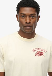 Camiseta de algodón amarilla clara con cuello redondo; presenta un gráfico rojo de "SUPERDRY" y una ilustración de un camión vintage en el área del pecho izquierdo.