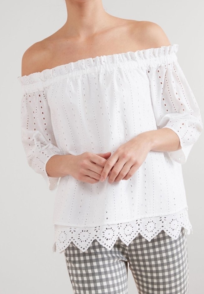 Femme portant un chemisier blanc à épaules dénudées en broderie anglaise avec un ourlet festonné et des manches trois-quarts, associé à un pantalon à carreaux gris et blanc.