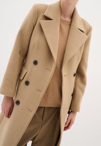 Beige vlněný kabát s podříznutým límcem, dlouhými rukávy a čtyřmi černými knoflíky. Nosený přes ribovaný béžový svetr a kalhoty.