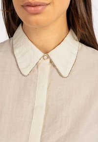Light beige linen shirt with a rounded collar, embellished with silver trim and a pearl button at the neck. Soft texture, casual design.