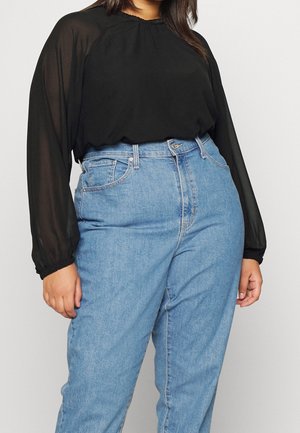 Kobieta ubrana w czarną, przejrzystą, długorękawową bluzkę wpuszczoną do jasnoniebieskich dżinsów z wysokim stanem, stojąca na jednolitym tle.