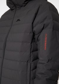 Svart vadderad jacka med dragkedja framtill, med horisontell sömnad och en logotyp på bröstet. Ärmen har röd text som accent.