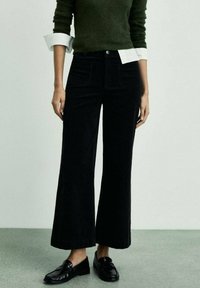 Pantaloni in corduroy neri a gamba larga con vita alta, tasche frontali e una superficie leggermente texturizzata. Indossati con un maglione lavorato a maglia verde.