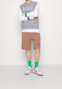 En person står med armarna i kors, klädd i en grå Nike-tröja, bruna shorts och gröna strumpor, tillsammans med vita sneakers med röda accenter.