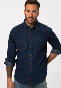 Donkerdenim knoopshirt met twee borstzakken, bruine knopen, opgerolde manchetten en een gebogen zoom. Beschikt over zichtbare stikselaccenten.