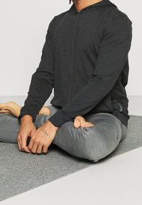 Chaqueta con capucha y cremallera en tela gris oscuro, combinada con pantalones tapered gris claro. Persona sentada sobre una estera gris, con las piernas cruzadas y las manos relajadas.