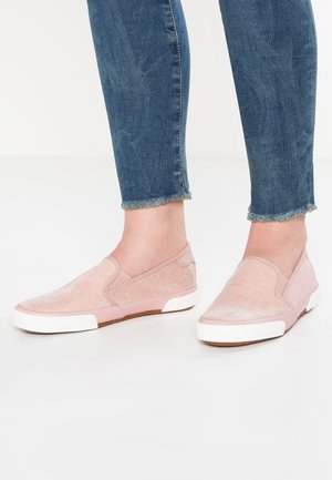Fødder iført lyserøde slip-on sneakers med hvide såler, kombineret med blå denimjeans med frynset kant.