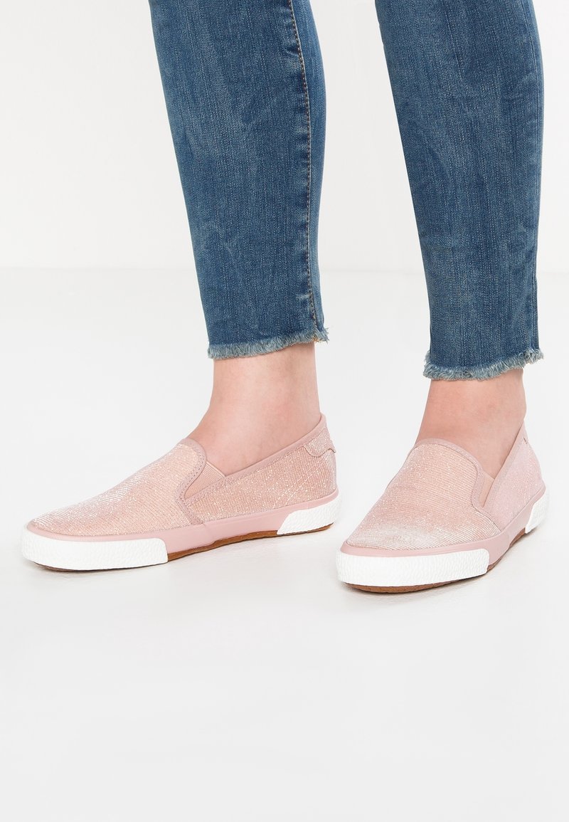 Pieds portant des baskets légères roses pâles sans lacets avec des semelles blanches, associés à un jean bleu en denim à ourlets effilochés.