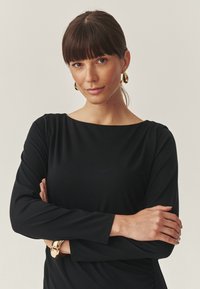 Top negru cu mânecă lungă, realizat dintr-un material moale, cu un decolteu larg. Cercei aurii și o brățară aurie completează ținuta.