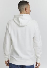 Witte pullover hoodie met een kangoeroezak, een gestructureerde stof, ribgebreide manchetten en een capuchon met trekkoord, gedragen door een persoon van achteren.