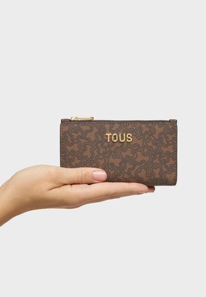 Mano che tiene una piccola pochette marrone a motivo TOUS con logo dorato su uno sfondo grigio chiaro uniforme.