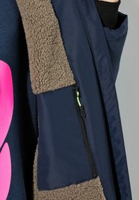 Chaqueta navy con un cuello de forro polar texturizado marrón y un bolsillo con cremallera en el lado izquierdo que tiene una pestaña de tirón en verde neón.