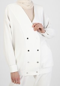 Witte geribbelde jas met een diepe V-hals, drukknoopjes en elastische manchetten, gecombineerd met een beige nekdoek daaronder.