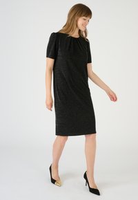 Vestito nero a maniche corte con una leggera lucentezza, caratterizzato da un collo plissettato e un taglio dritto. Indossato con tacchi a punta neri e dorati.