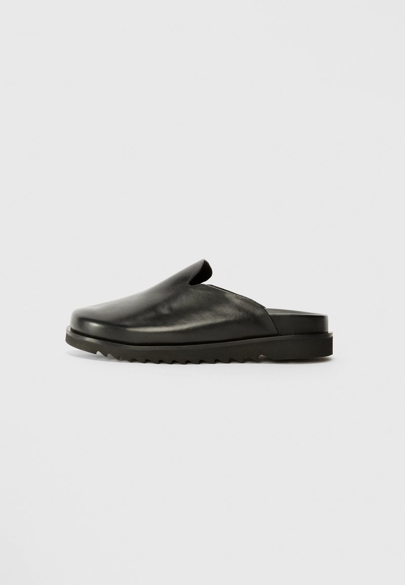 Black leather slip-on shoes with a smooth finish, rounded toe, and textured rubber sole featuring a zigzag pattern. Minimalist design.