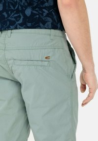 Hellgrüne Shorts aus leichtem Stoff. Mit einer Gesäßtasche mit braunen Nähten und einem kleinen Logo-Akzent auf der Tasche.