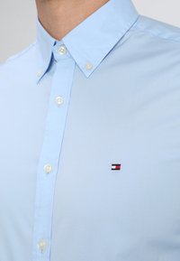 Camisa de botones de color azul claro con una textura suave, que presenta un pequeño logotipo bordado en rojo, blanco y azul en el pecho.