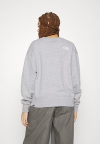 Felpa grigia con un taglio rilassato, polsini e orlo a coste. Presenta il logo North Face in bianco sulla parte superiore della schiena. Il materiale sembra morbido.