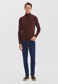 Brązowy sweter z golfem, dopasowany krój, w zestawie z ciemnoniebieskimi dżinsami. Model stoi w zrelaksowanej pozycji, ma na sobie brązowe buty z beżowymi podeszwami.
