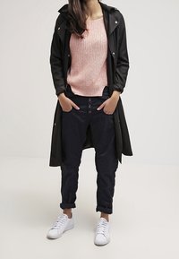 Czarny płaszcz trench, jasnoróżowy sweter z dzianiny, ciemnoniebieskie spodnie z guzikami i białe sneakersy. Prosty, codzienny strój z różnorodnymi teksturami.