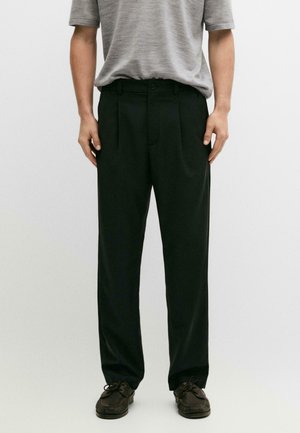 Man wearing loose black trousers, gray short-sleeve shirt tucked in, and dark brown lace-up shoes, standing against plain background.