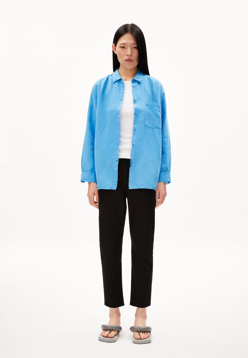 Hellblaues Button-down-Hemd mit einer einzelnen Brusttasche, weißes Tanktop, schwarze schmal zulaufende Hose und graue flache Sandalen mit Riemendetails.