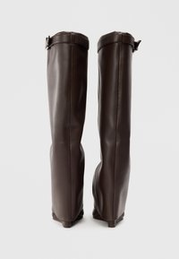 Bottes cuissardes en cuir marron au design élégant, avec un haut roulé et une sangle latérale pour le maintien.