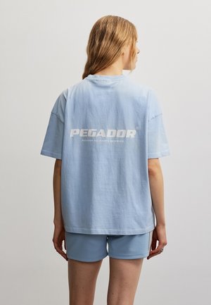 Personne aux cheveux châtain clair portant un t-shirt bleu clair oversize avec le texte « PEGADOR » et un short assorti, vue de dos sur un fond uni.