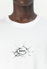 Fehér pamut póló, fekete grafikával, amelyen a "BOSS" szó látható egy repedt felületi mintában. Kerek nyakkivágás rövid ujjú.