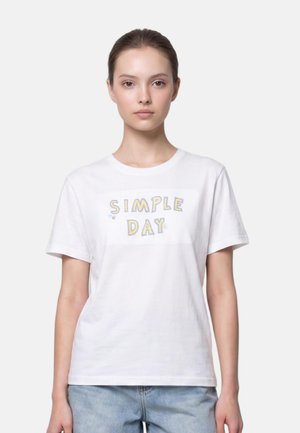 Jeune femme aux cheveux bruns portant un t-shirt blanc avec le texte "SIMPLE DAY" et un jean bleu clair, debout devant un fond uni.