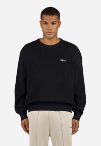 Sort ribbet sweater med rund hals, løs pasform og lange ærmer. Har hvid logo broderi på brystet. Bæres med beige bukser.