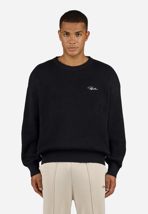 Black ribbed sweater with round neck, loose fit, and long sleeves. Features white logo embroidery on the chest. Worn with beige trousers.
