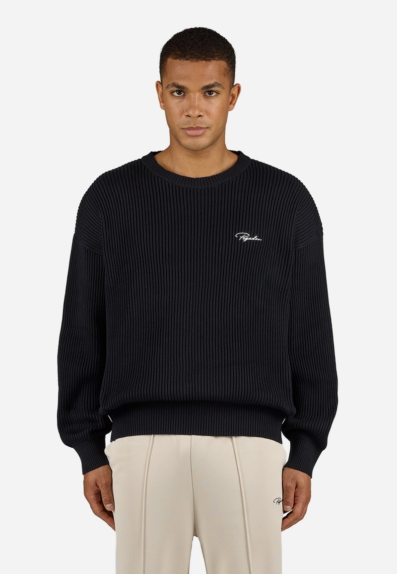 Sort ribbet sweater med rund hals, løs pasform og lange ærmer. Har hvid logo broderi på brystet. Bæres med beige bukser.