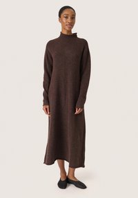 Braunes, mittellanges Strickkleid mit hohem Kragen und langen Ärmeln, aus einem strukturierten Stoff gefertigt. Das Model kombinierte es mit schwarzen Ballerinas.