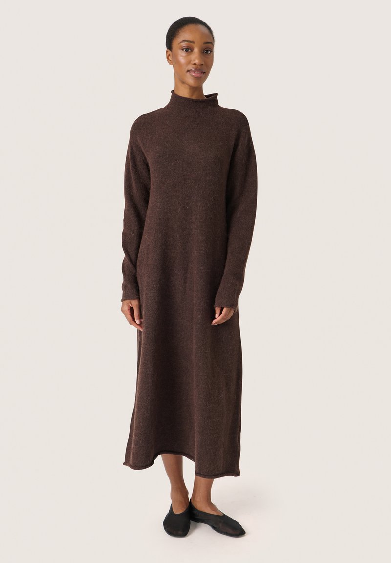 Braunes, mittellanges Strickkleid mit hohem Kragen und langen Ärmeln, aus einem strukturierten Stoff gefertigt. Das Model kombinierte es mit schwarzen Ballerinas.