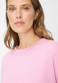 Pull rose clair avec un col rond et une coupe légèrement ample. Texture lisse et design minimaliste sans motifs ni accessoires visibles.