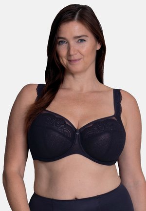 Frau mit langen braunen Haaren, die einen schwarzen Spitzen-BH mit Bügeln trägt, lächelnd vor einfachem hellem Hintergrund.