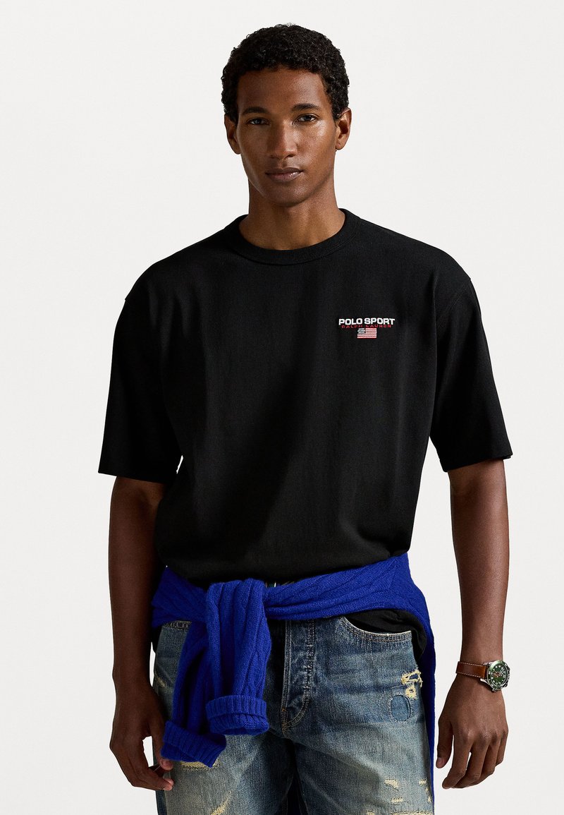 Camiseta de algodón negra de manga corta con el logo "POLO SPORT" en rojo. Suéter azul atado a la cintura, jeans de mezclilla desgastados y reloj.