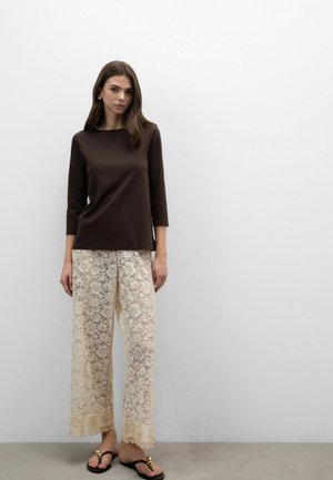 Frau steht an einer weißen Wand und trägt ein dunkelbraunes Oberteil mit Dreiviertelärmeln, cremefarbene, breite Hose mit floralem Spitzenmuster und schwarze Sandalen mit goldenen Akzenten.