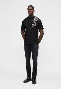Homme debout, les mains dans le dos, portant un t-shirt noir avec un motif de serpent bleu sur l'épaule, un jean noir et des chaussures en cuir noires.