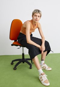 Pantaloni neri a vita alta, top arancione, e sneakers bianche con accenti marroni, seduto su una sedia a rotelle nera su un tappeto verde.