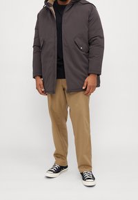 Grå vadderad jacka med huva, dragkedjestängning och sidofickor, i kombination med beige byxor och svarta sneakers med vita accenter.