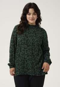 Green long-sleeve top with black abstract print, featuring a high neckline and relaxed fit. Soft fabric with a smooth texture.