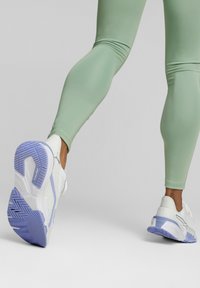 Hvid atletiksko med en struktureret overdel og lilla sål, kombineret med tætsiddende lysegrønne leggings. Fokus på stil og formdetaljer.