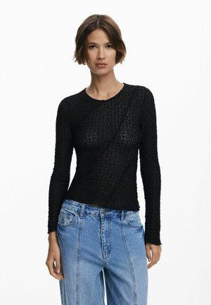 Maglia nera a maniche lunghe con design tessuto e strutturato; scollatura rotonda e vestibilità slim. Abbinata a jeans a vita alta larghi di colore blu chiaro.
