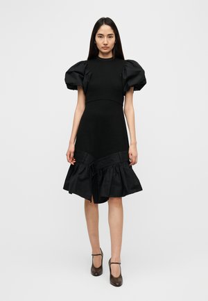 Robe noire à manches bouffantes avec corsage cintré et ourlet à volants. Fabriquée dans un tissu extensible, dotée d'une finition texturée et d'un détail cordon de serrage.